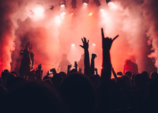 Live concert stage with band performing under red lights and smoke, crowd with hands raised