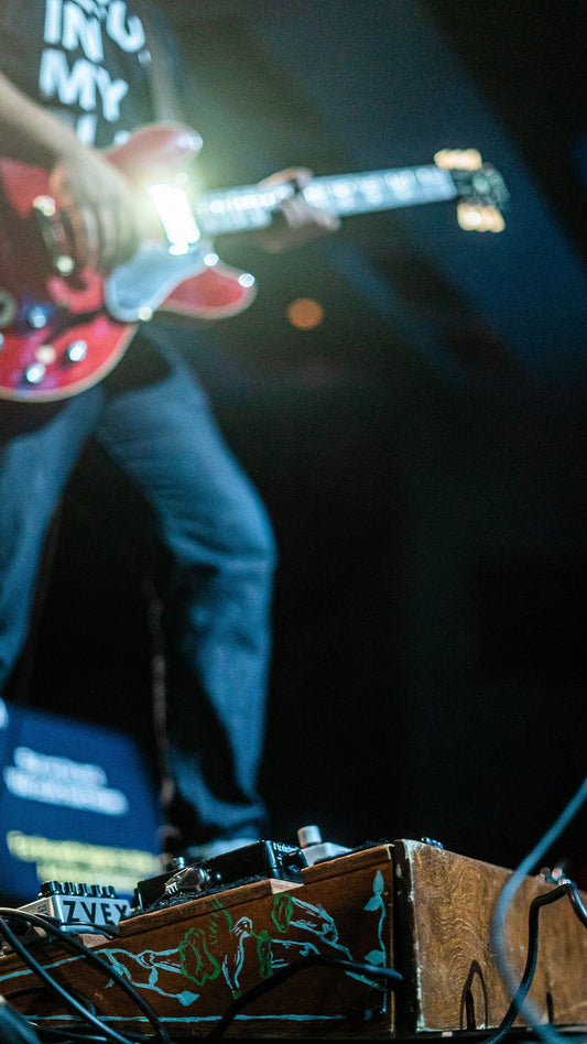 Guitarist performing on stage with pedalboard and effects pedals in the foreground under concert lighting